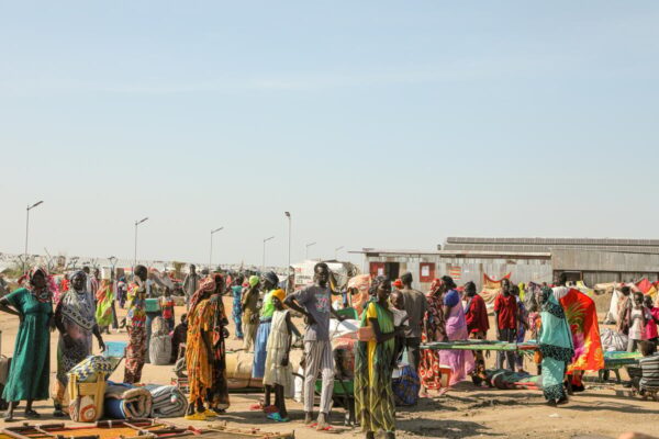 A crowd of refugees - many carrying their belongings - stands on a dusty open field in the Renk transit centre for refugees arriving into South Sudan from Sudan.