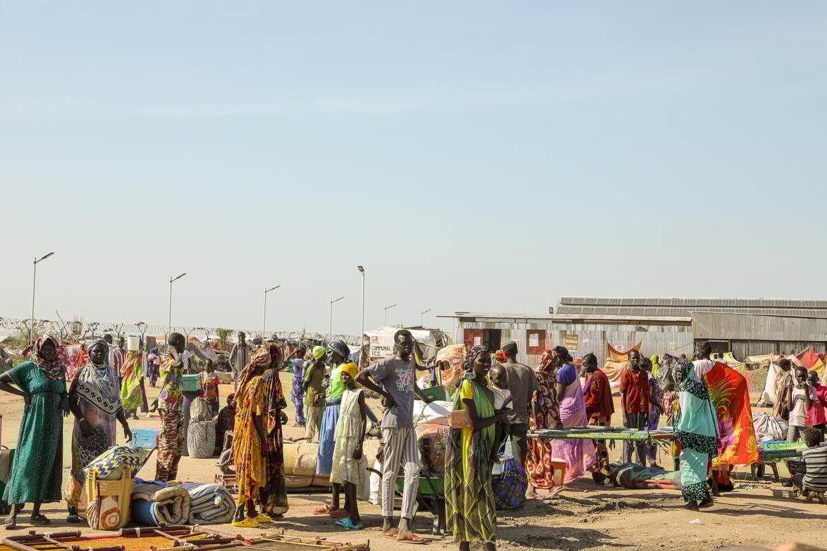 A crowd of refugees - many carrying their belongings - stands on a dusty open field in the Renk transit centre for refugees arriving into South Sudan from Sudan.