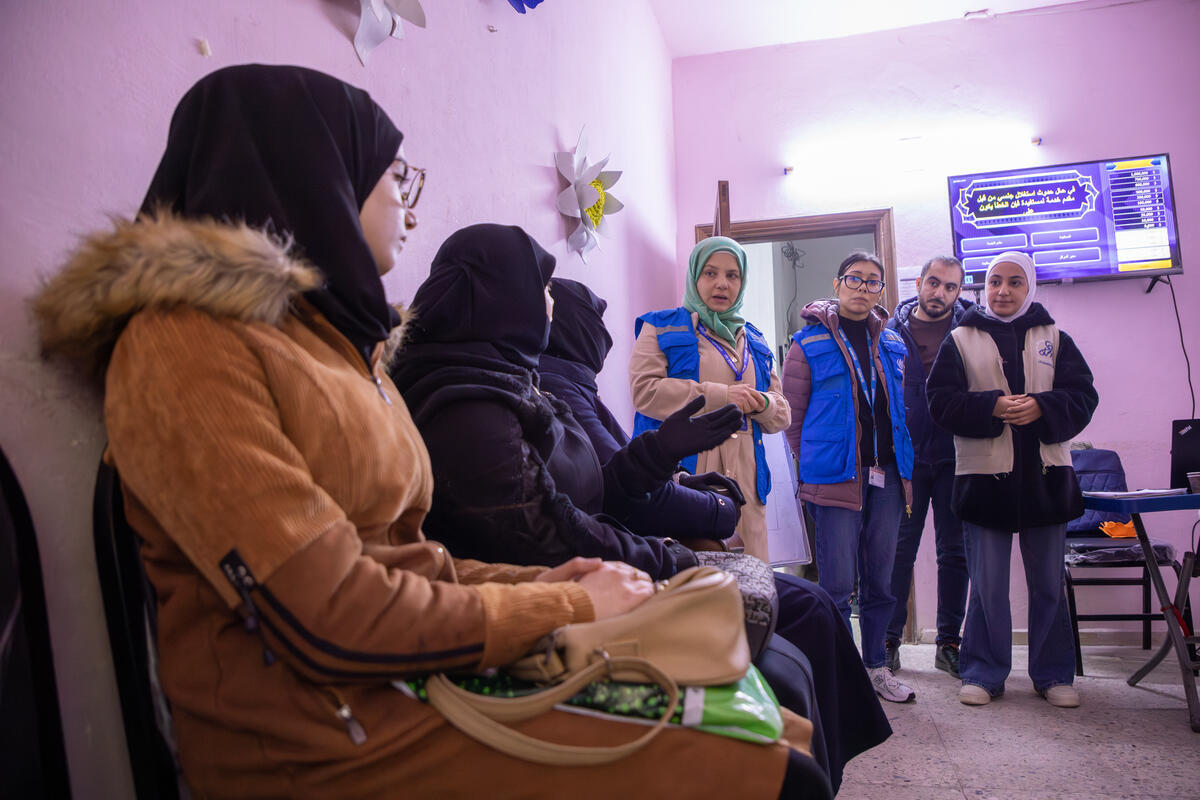 Un groupe de femmes portant des hijabs noirs et des manteaux d'hiver est assis dans la salle d'attente d'un centre communautaire, tandis que trois autres femmes et un homme se tenant près de la porte et portant des vestes bleues du HCR leur parlent.