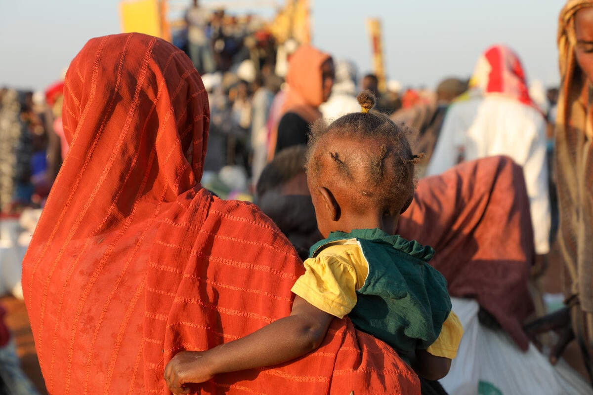 A photo of a woman with her back to the camera. She wears an orange headscarf over her head and shoulders while holding in her arms a small girl in a bright yellow dress and her hair braded into short braids. 