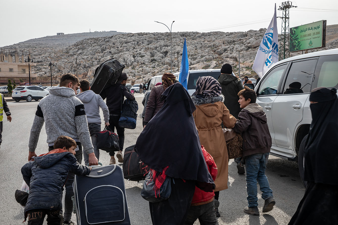Une grande famille de réfugiés syriens avec des enfants et des valises marche sur une route tandis qu'un HCR flotte au vent à l'arrière-plan.