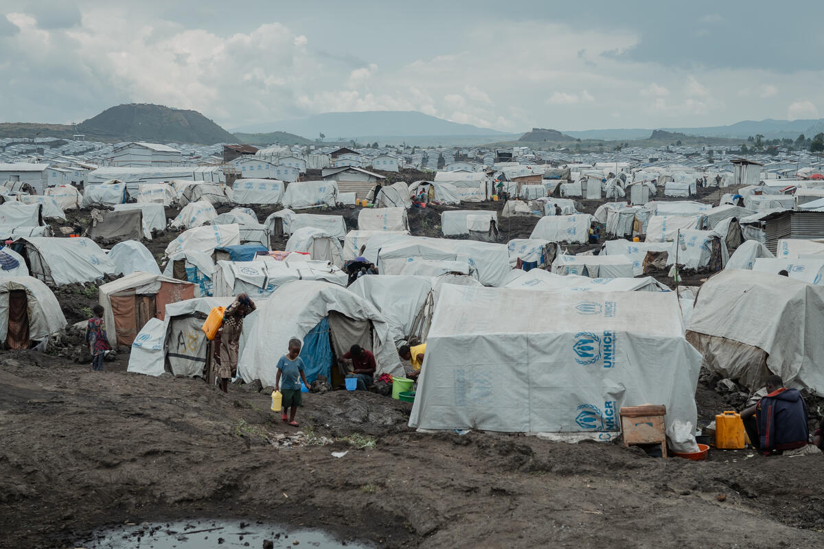 Un jeune enfant marche sur un sol boueux devant des centaines de tentes blanches du HCR installées dans une plaine où l'on aperçoit des montagnes au loin.
