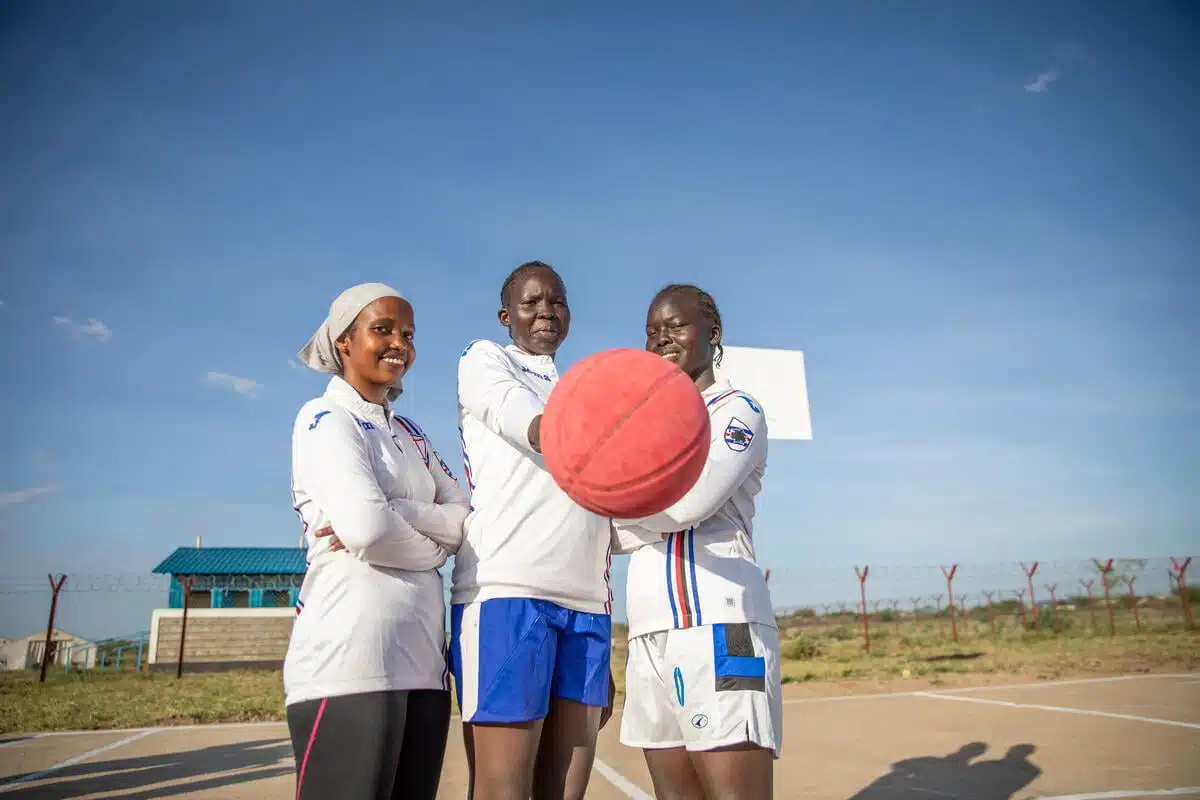 Three African American women smiling on a sports court, dressed in white uniforms and holding a basketball.