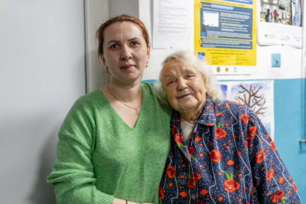 Two women, a middle aged woman dressed in a green v-neck sweater and an older, white-haired woman dressed in a floral pattern dress, hug each other and look smiling at the camera.