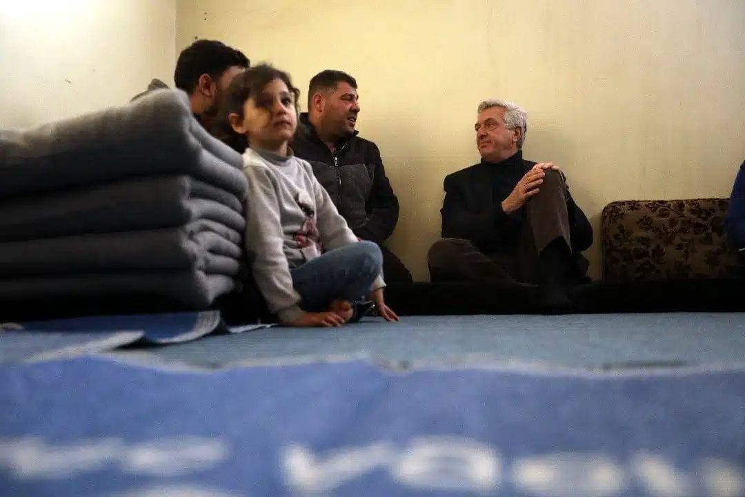 A little girl sits cross-legged on the floor next to a stack of folded blankets as three older pictured in the background men converse with each other. 