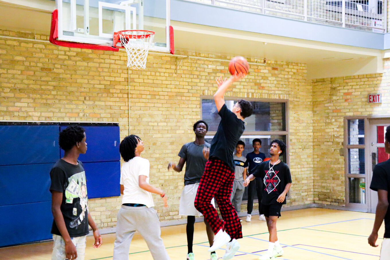 A young boy wearing a black t-shirt and black and red checkered pants jumps to throw a basketball into the hoop. 