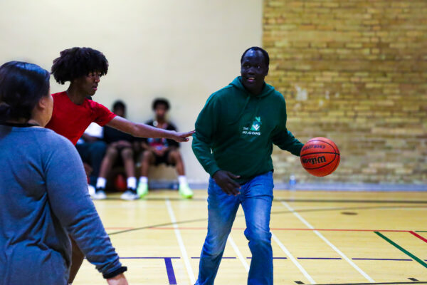 Photo of a man wearing a green hoodie and blue jeans hold a basketball and runs in a basketball court, with a young boy in a red t-shirt reaching for the ball to his left hand side.