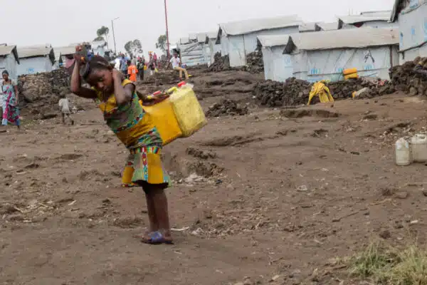 A young girl carries two large yellow plastic water jars on her back that are strapped to her forehead . In the background a row of white shelters