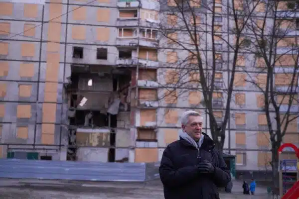 UN High Commissioner for Refugees Filippo Grandi stands in front of a damaged multi-storey building with damage from bombing visible on the facade.