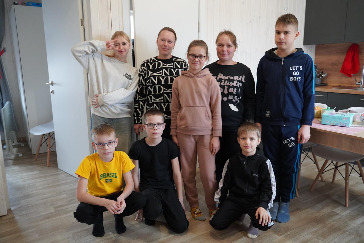 A family of eight members smiles at the camera. There are three small children, one teenager, two teenage girls, and two women standing behind them. They are in their new home.