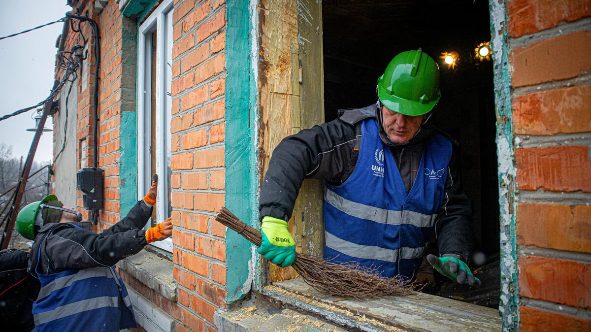 A man wearing a UNHCR vest and a green helmet is cleaning the edge of a window with a small wooden broom.