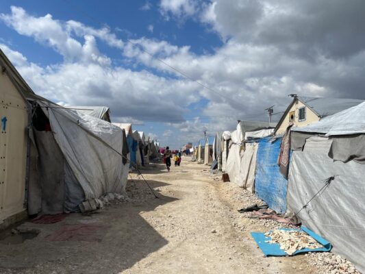 A group of children are seen walking on a narrow road lined with rows of tents and temporary shelters in a camp for internally displaced people.