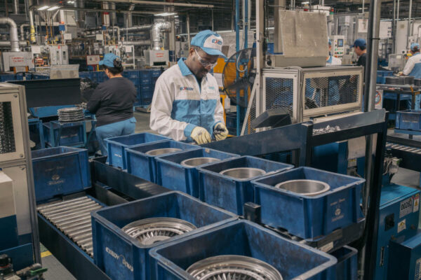 A black man dressed in white and blue overalls and wearing same colour baseball cap and safety glasses stands at an assembly line on a factory floor filled with machinery.