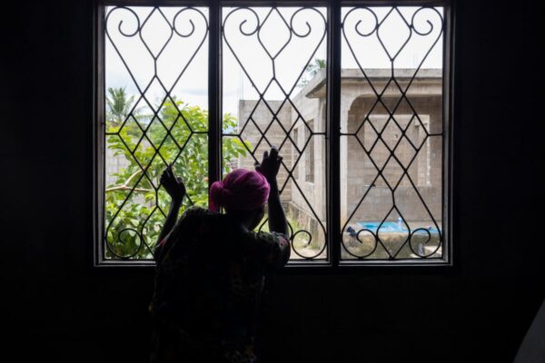 A haunting photo of a woman, with only her silhouette visible, looking out from a window onto a yard with some trees and unfinished houses under construction.