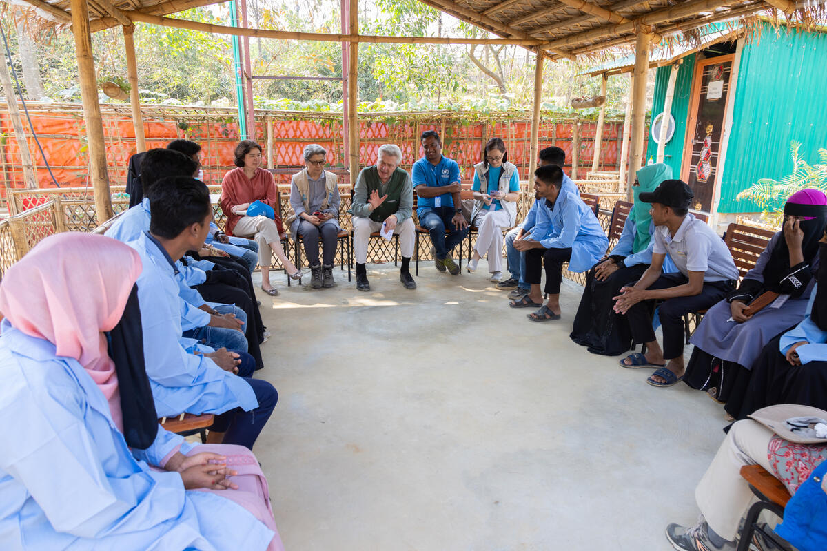 The High Commissioner is seated in a conference, surrounded by people who are listening to him attentively. The event is taking place in an open space with trees in the background.