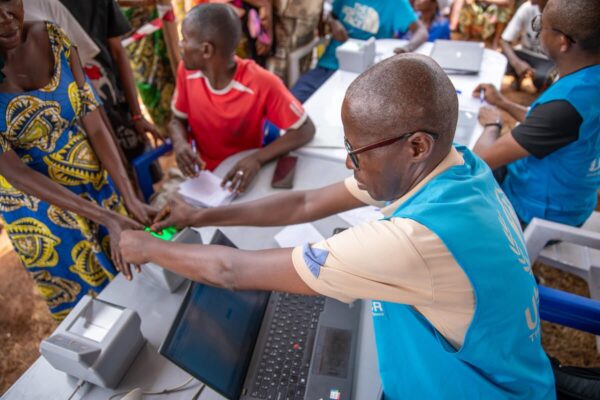 A middle-aged and bald black man wearing spectacles and a light blue UNHCR visibility vest helps a woman wearing a colourful dress get her fingerprints scanned at a busy registration centre set up by UNHCR.