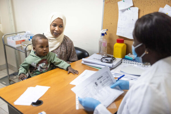 A Sudanese woman with small child on her lap sits across the table from a physician in a doctor's office.