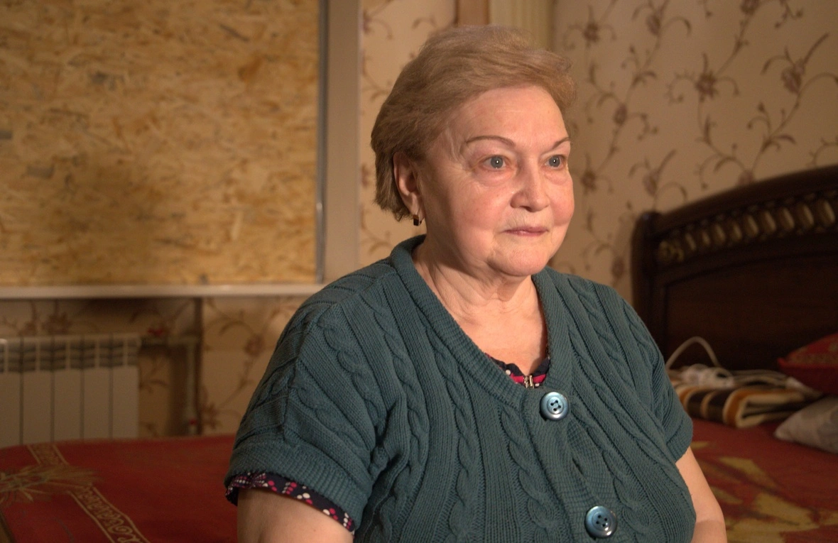 An elderly woman is calmly staring away from the camera. She is wearing a green short-sleeved sweater. She has short blonde hair and green eyes.