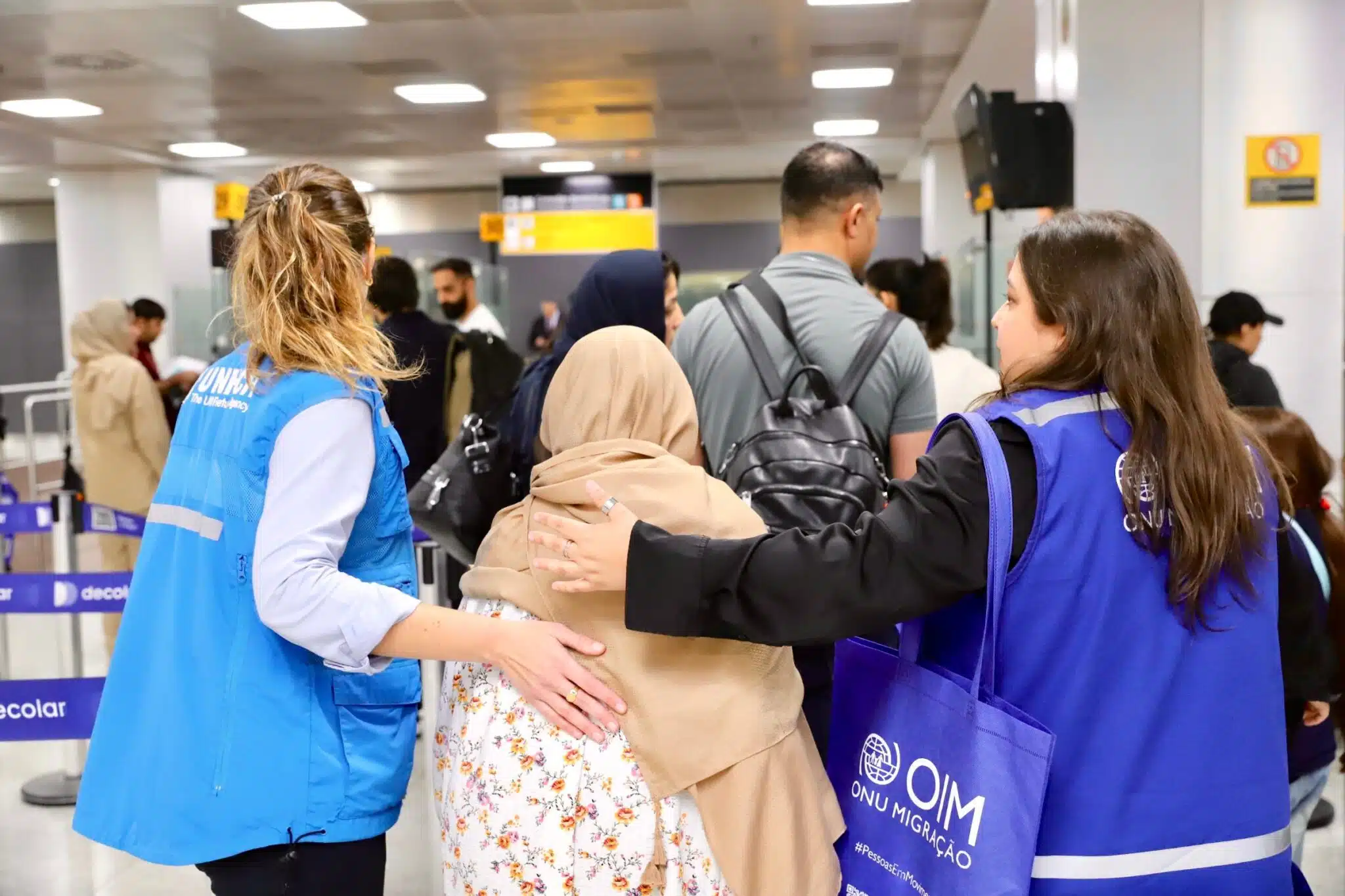 An female Afghan refugee dressed in a beige hijab is accompanied by two women on her left right sides, one wearing a UNHCR vest and the other an IOM vest in what looks like a busy airport arrival hall.