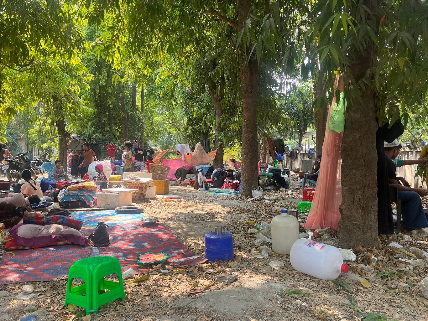 A group of people sits under the trees will all their belongings strewn around.
