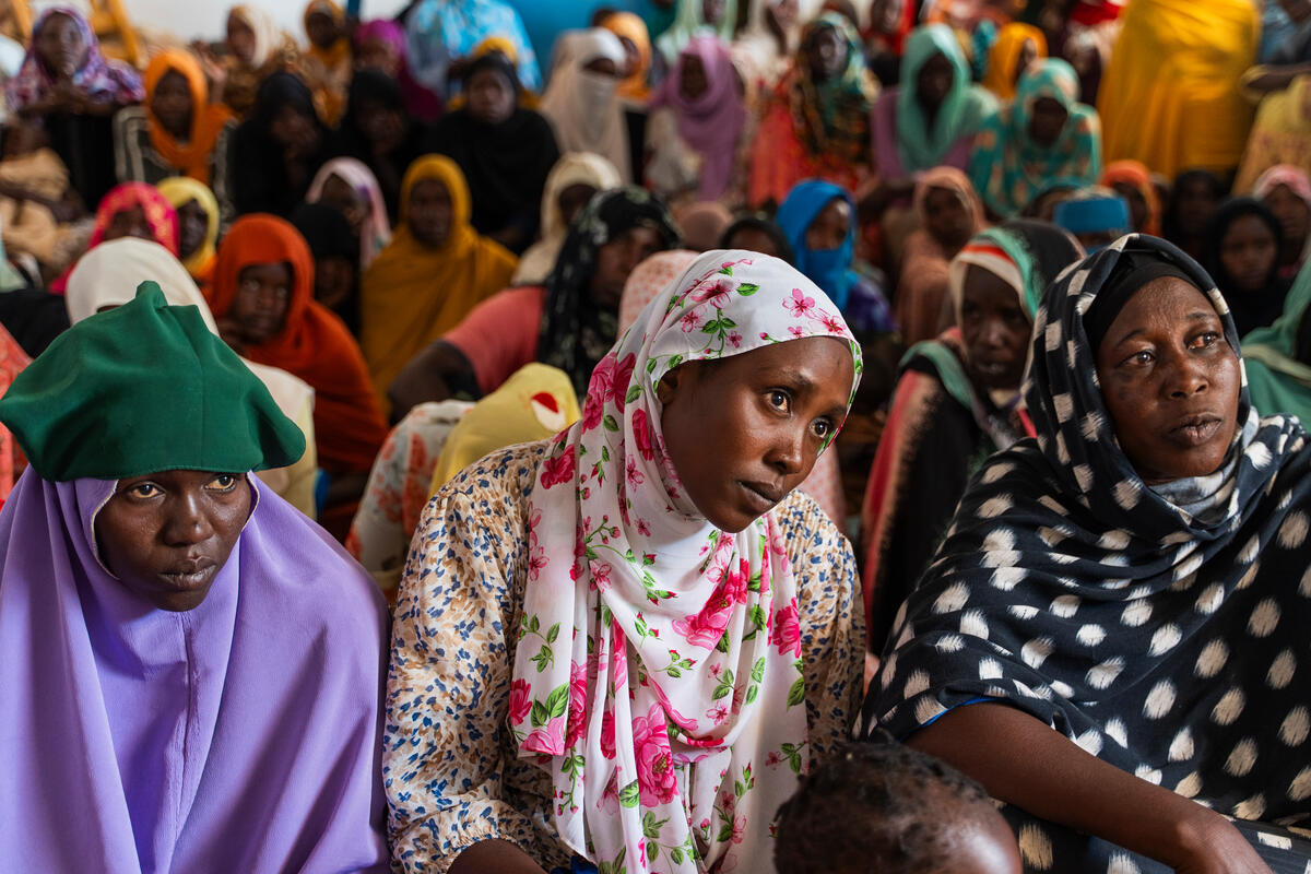 Several women are seated, listening attentively. They are all wearing colorful clothes paired with scarves on their heads, creating a beautiful contrast.