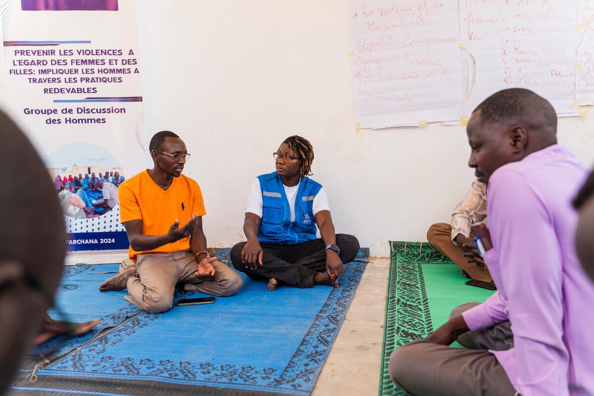 Three people are seated on the floor, chatting with each other. Behind them, there's a poster about violence against women and children.