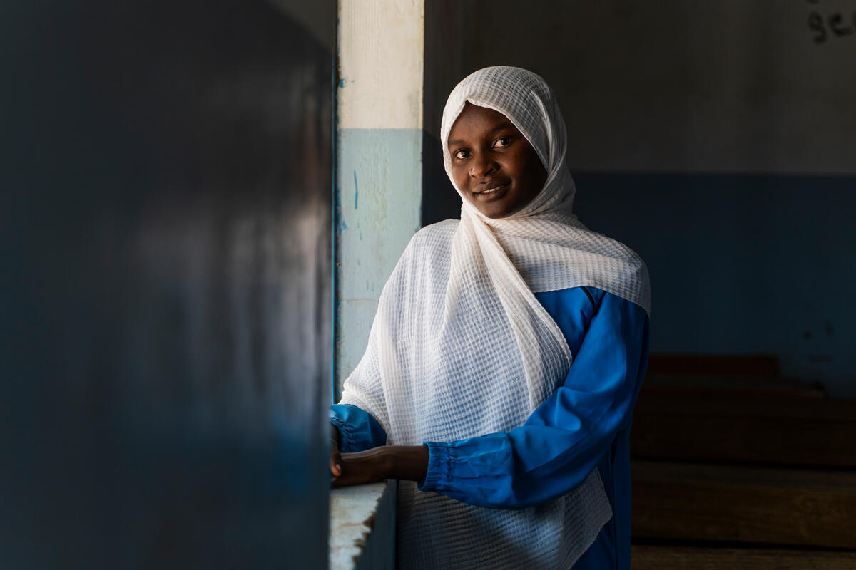 A young girl looks at the camera with a kind and smiling expression. She’s wearing a blue tunic and a beautiful white scarf on her head that drapes over her shoulders down to her waist.