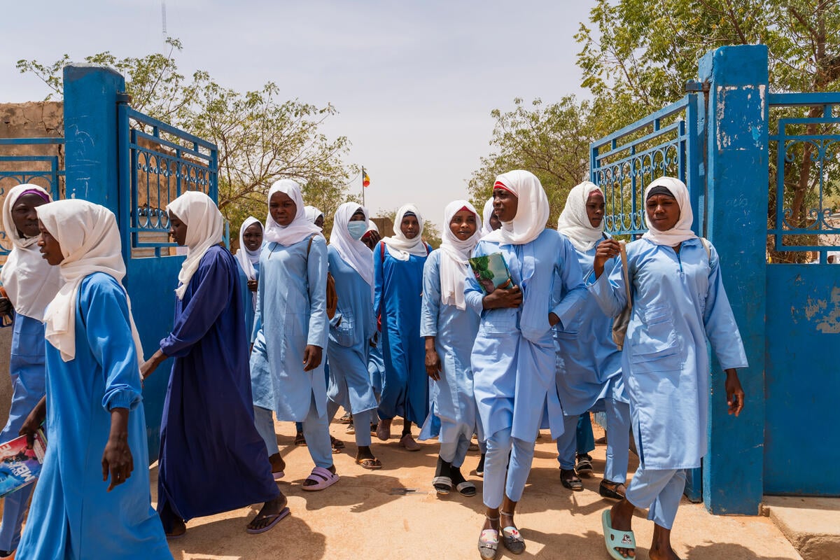 Several young girls are walking out of a school building. They are all wearing blue tunics and white scarves on their heads, and some are holding books.