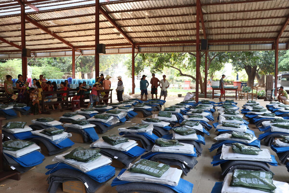 Rows up rows of neatly arranged relief items, including blankets and tarps are arranged on the floor in a large structure under a canopy roof as people in the background wait in line.