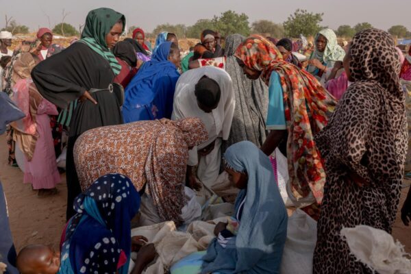 A group of Sudanese refugees in a crowded field gather around sacs of food on the ground as they measure and pack food rations in the sacs.