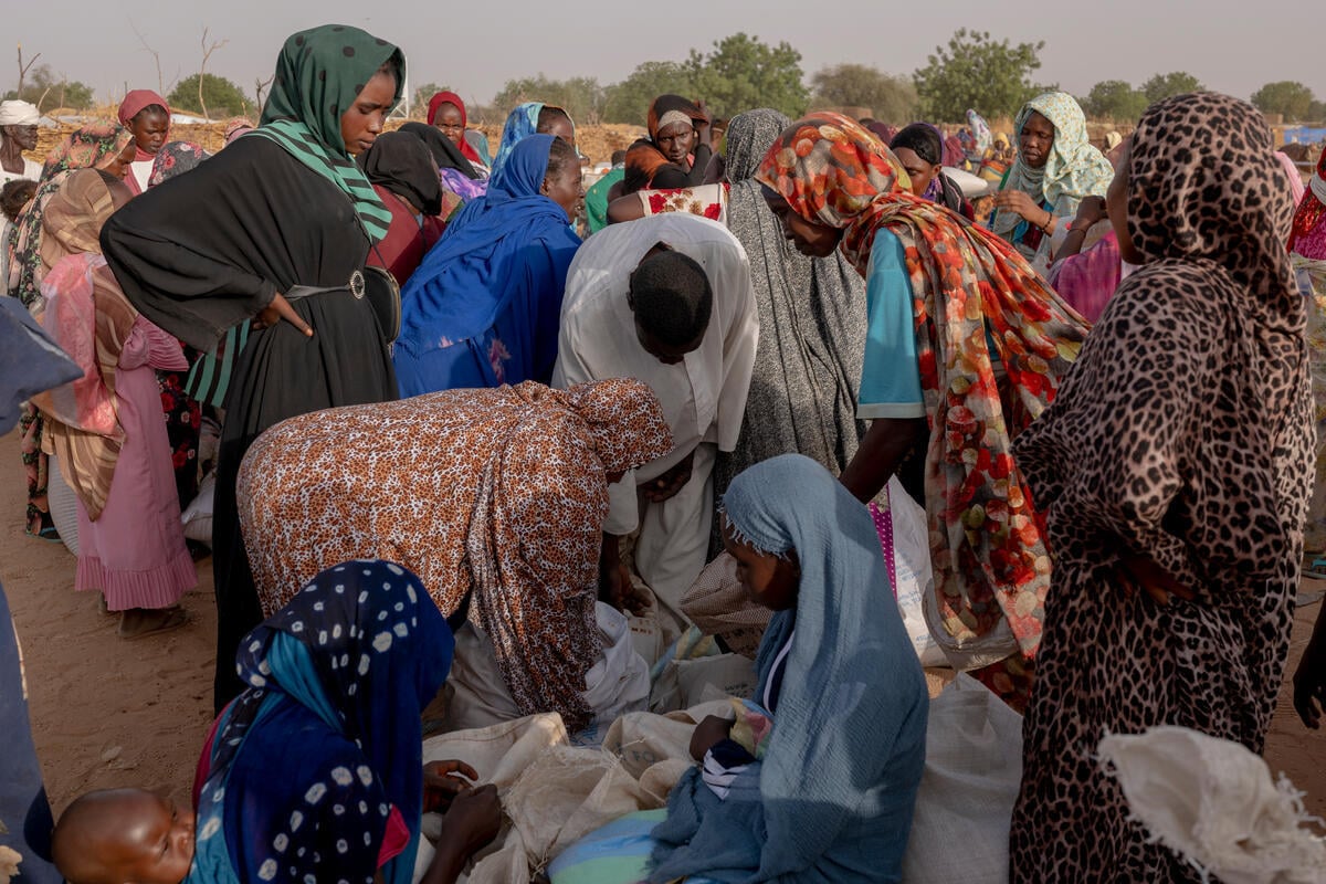 A group of Sudanese refugees in a crowded field gather around sacs of food on the ground as they measure and pack food rations in the sacs.
