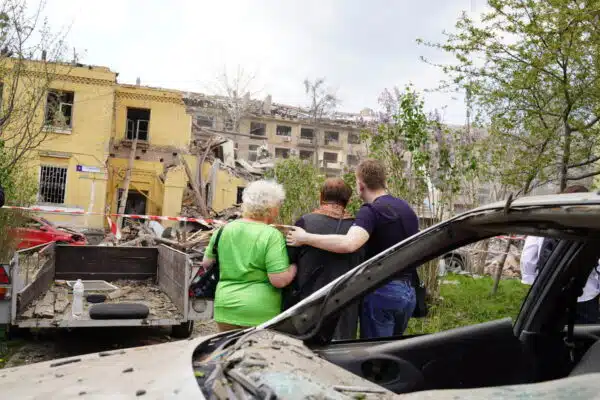Two older women and a younger man seen from behind are hugging each other as they look upon a severely damaged yellow brick building in the background. A charred remains of a car can be seen in the foreground.