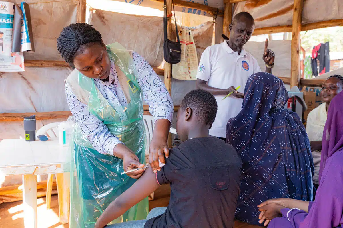 A black female nurse wearing a transparent plastic protective gown over her clothes administers a shot of vaccine in the upper arm of a teenage boy sitting in front of her inside a makeshift structure, while two other women sitting next to him speak with a male health worker.