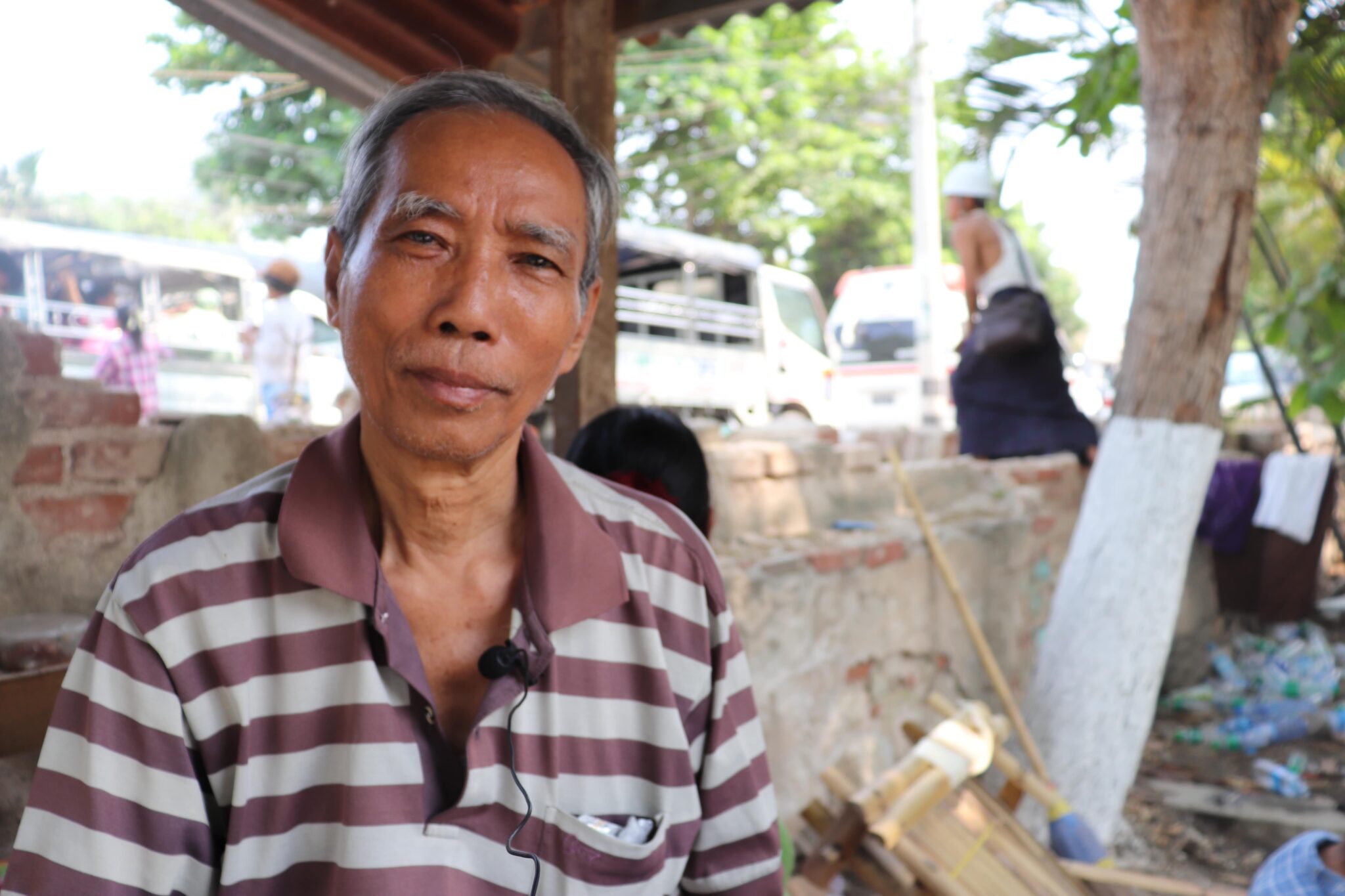An elderly man who is wearing a shirt with brown stripes and is seated at an open space where there's a tree behind him, smiles at the camera.