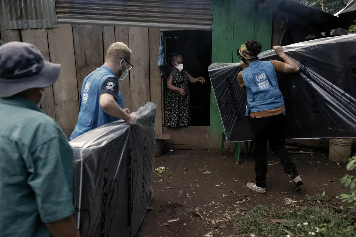 A male and a female UNHCR staff members wearing blue UNHCR visibility vests and medical masks carry new mattresses wrapped in plastic as an elderly woman greets them at the entrance of a house built from rough wooden planks and corrugated iron sheets.