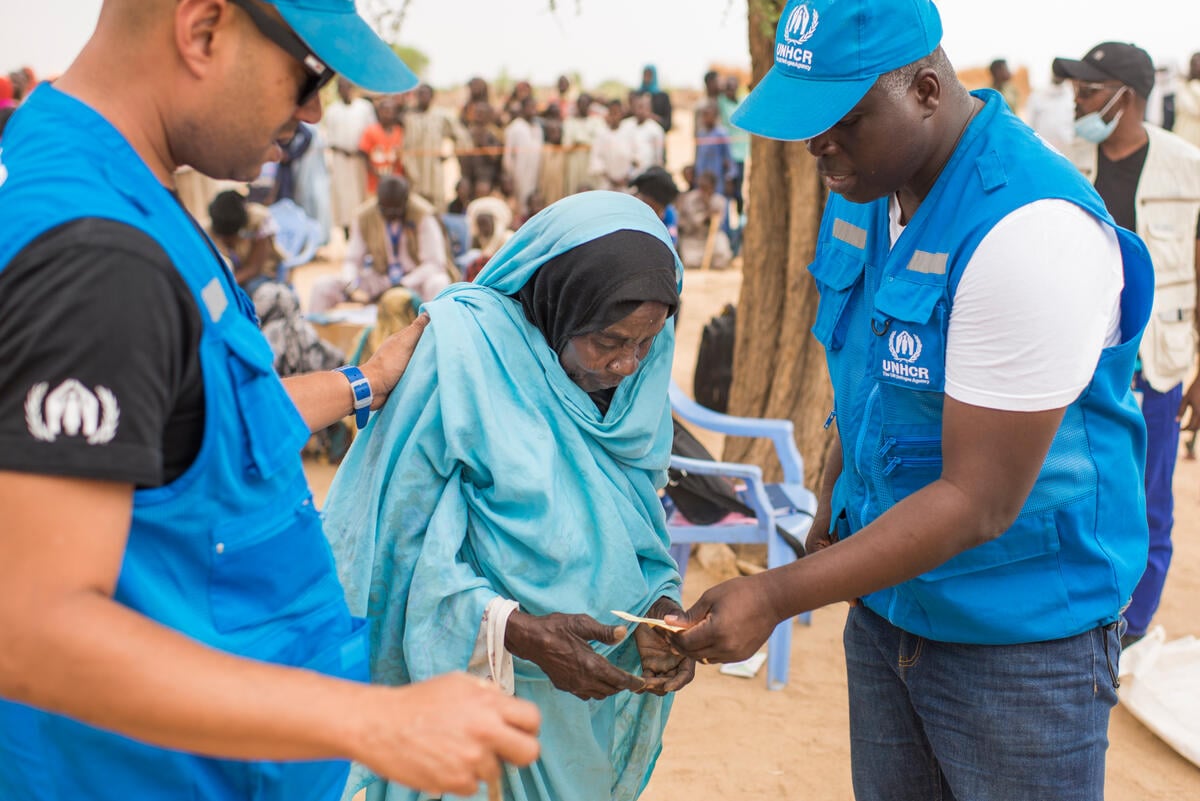 Two UNHCR men are helping an elderly woman as she hands over some papers.