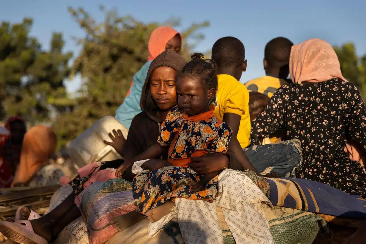 A young person dressed in a brown hoodie holds in their arms a small girl as they seat on top of a horse-drawn cart with other members of their family.