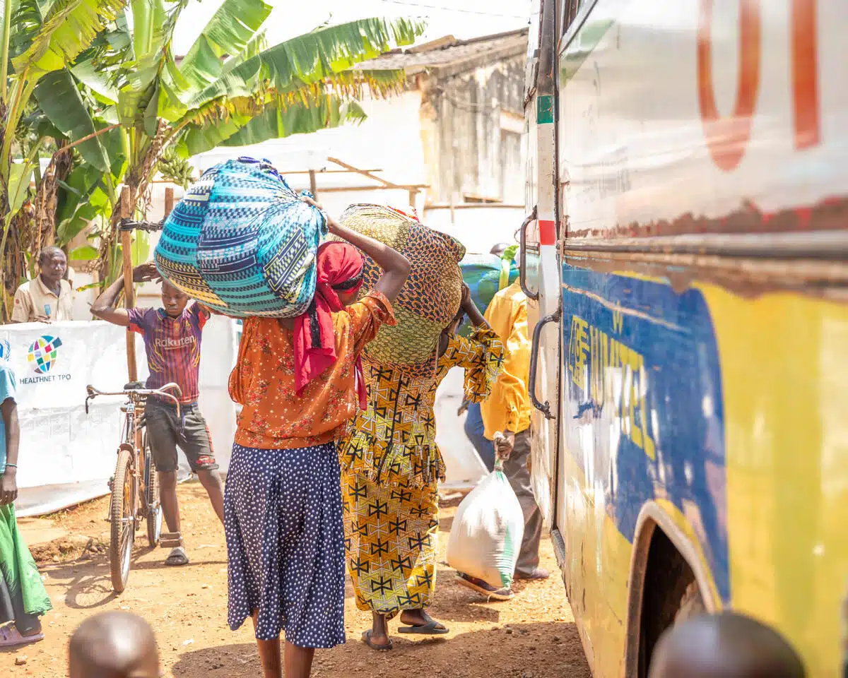 Two women carry large bundles of belongings on their heads as they load their luggage onto a bus.