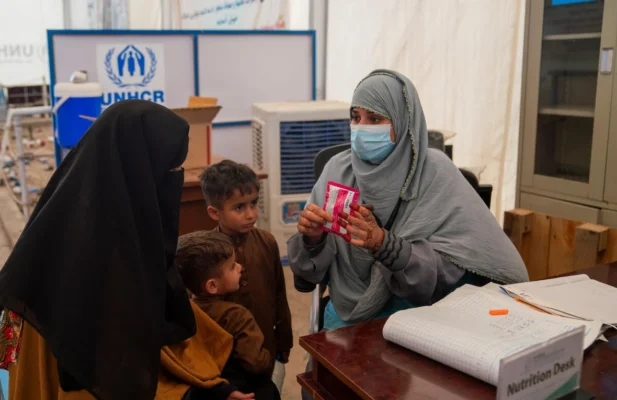 An Afghan woman in a black chador head covering with two little boys in tow sits across the table from a UNHCR nurse wearing a surgical mask and a light blue headscarf and holding a package of medication in her hands adorned with henna decorations.