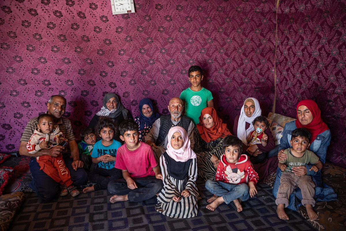 A family of children and adults is sitting on a carpet, looking at the camera. They are wearing colorful clothes and appear serious.