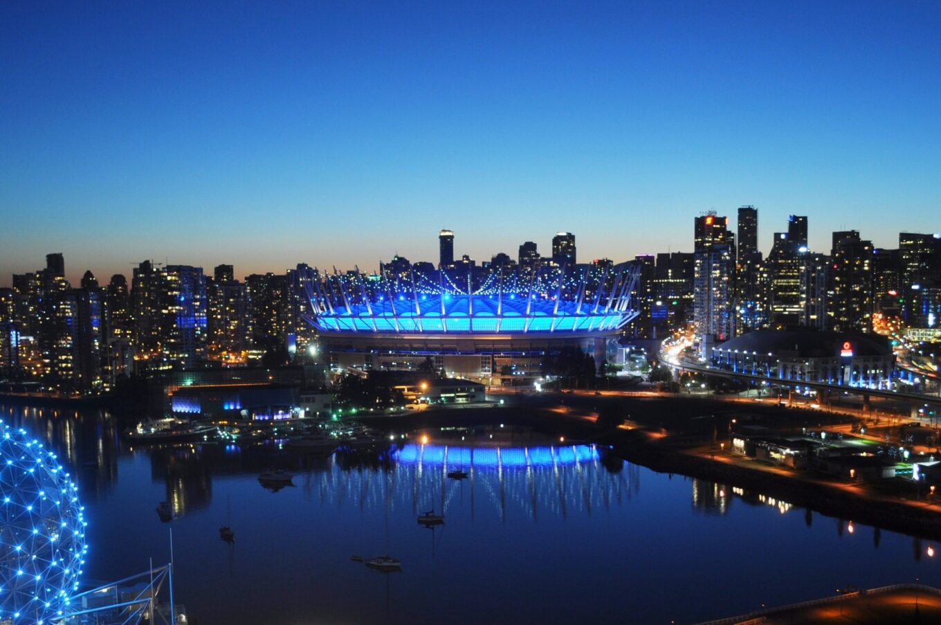 BC Place in Vancouver illuminated blue at night, with its roof glowing and reflecting in the nearby water against the city skyline.