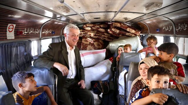 Un homme d'âge moyen souriant, vêtu d'un costume marron et d'une chemise blanche, se tient debout dans un bus, entouré d'enfants et de femmes assis autour de lui, et de quelques matelas empilés à l'arrière.