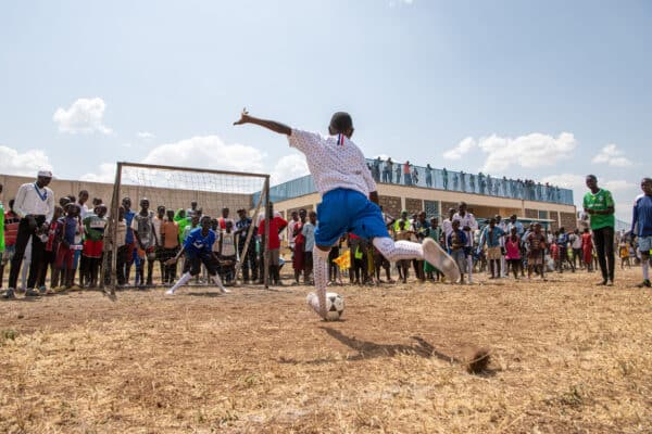A young girl wearing a white t-shirt and blue shorts kicks a soccer ball towards a soccer net. Behind the net onlookers watch her play.