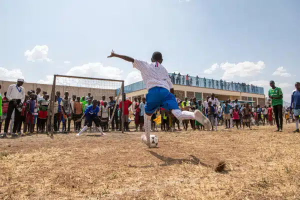 A young girl wearing a white t-shirt and blue shorts kicks a soccer ball towards a soccer net. Behind the net onlookers watch her play.