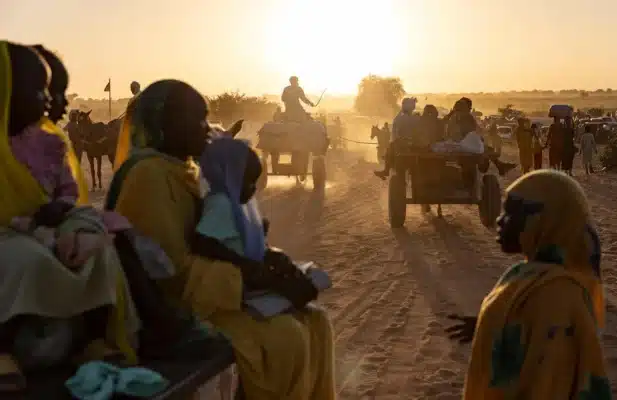 A group of women and children is seen seated on horse-drawn carts on a dusty desert road filled with other people and carts as the sun sets in the background.