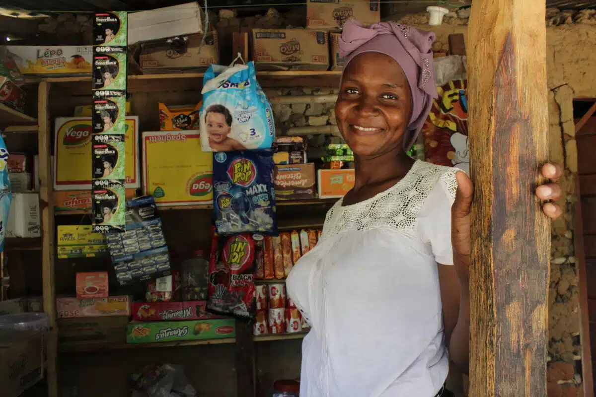 A woman stands at the entrance of a convenience store, wearing a brown scarf and a white blouse. She smiles warmly at the camera.