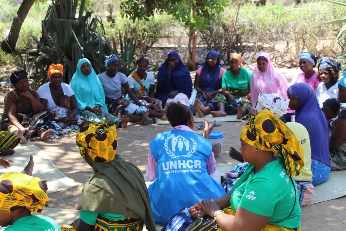 A group of women is seated in a circle in an open space. They are all wearing colorful scarves and outfits