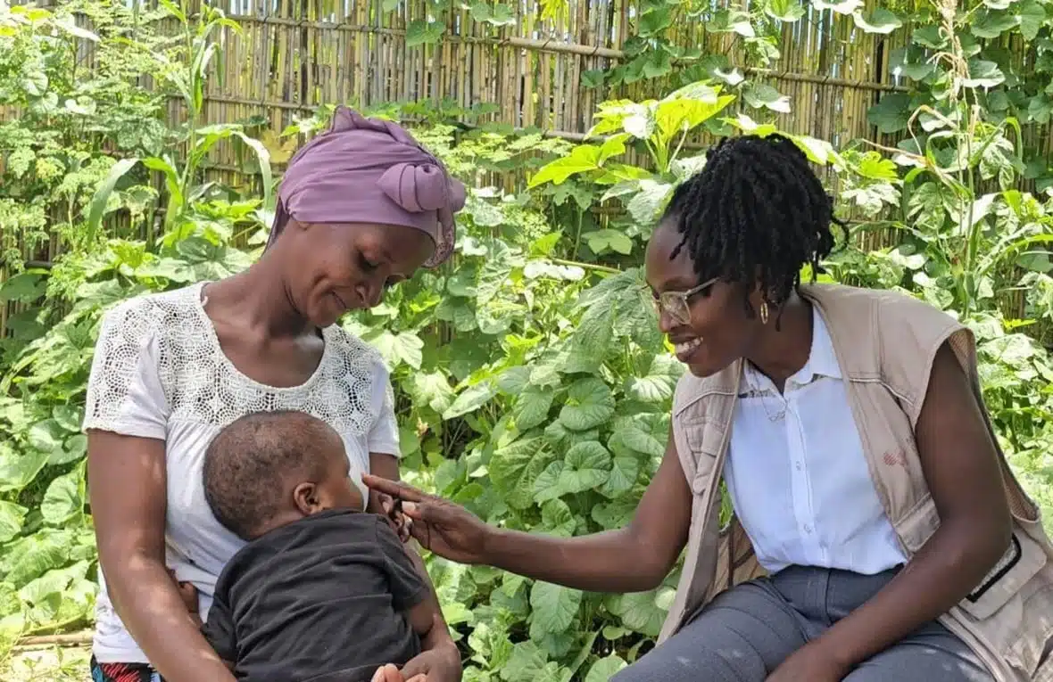 Two women are seated next to each other in a garden. One of them is holding a baby in her lap, and the other is gently caressing the baby’s face while smiling warmly