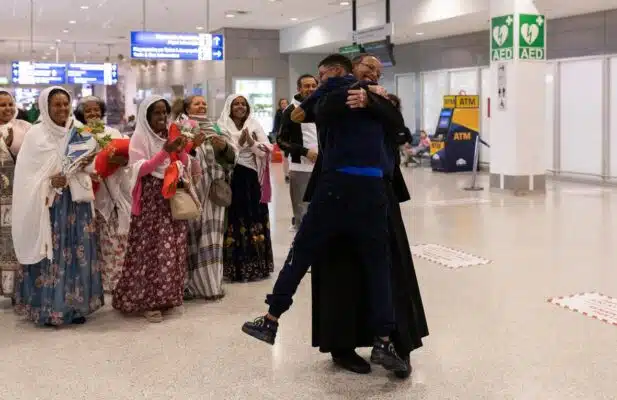 Two men are hugging joyfully at the arrival hall of an airport as a group of women in colourful dresses watch smiling.