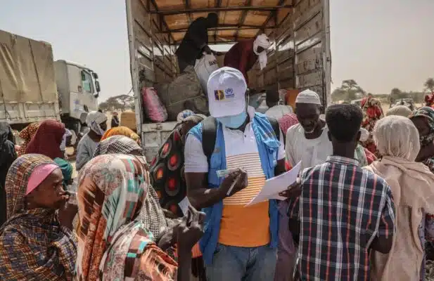 A male UNHCR staff member wearing a white baseball cap and a blue UNHCR vest over a white and orange t-shirt looks at an identity card of a Sudanese refugees while surrounded by several women and men. In the background workers are unloading a truck with humanitarian aid.